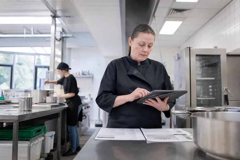 Eine Küchenleitung vergleicht Rezepturangaben auf einem Tablet mit einem Wochenplan